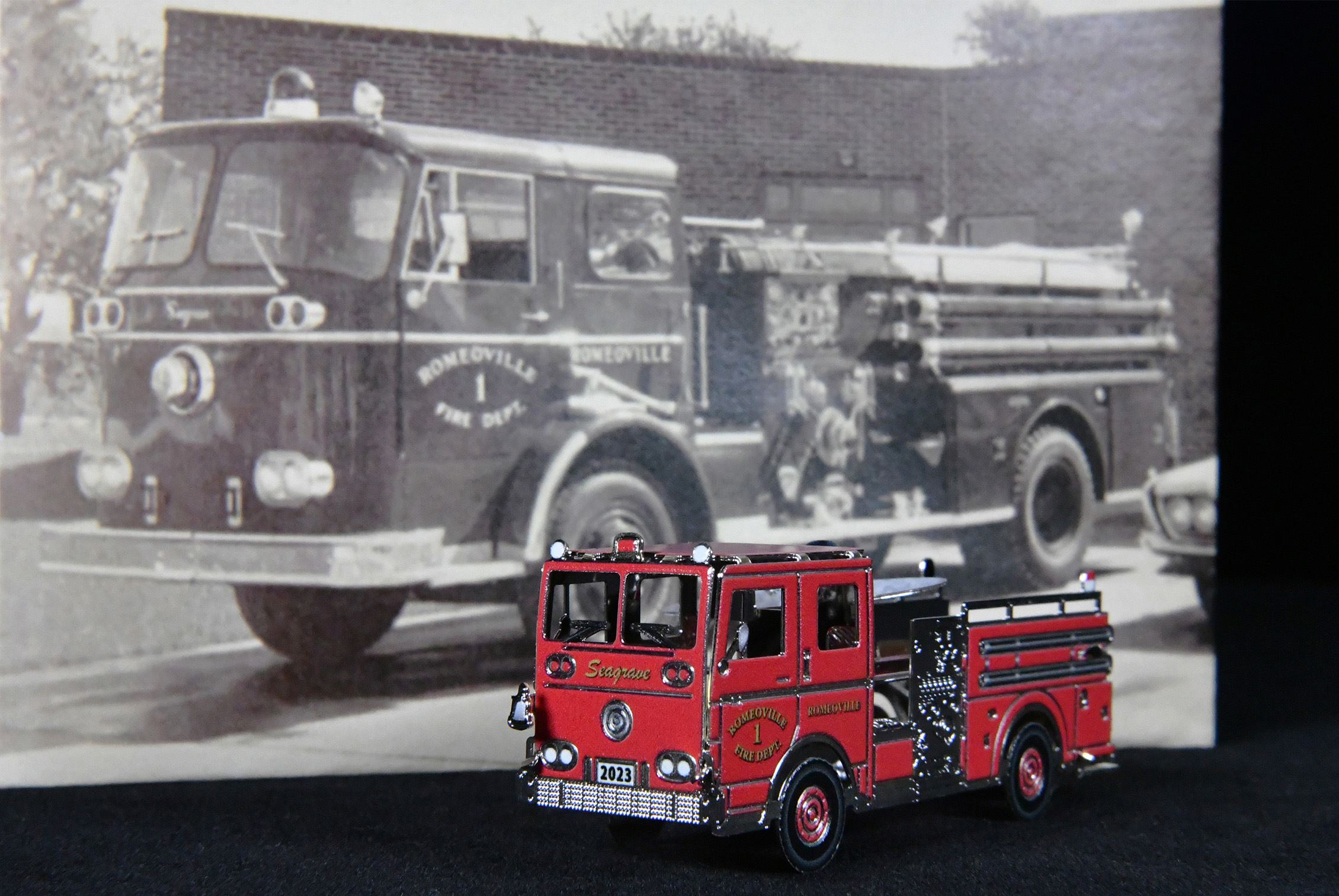 An ornament shaped like a red firetruck sits in front of a photo of an old firetruck.